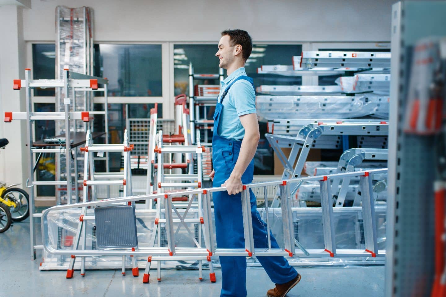 Männlicher Angestellter in Uniform hält neue Aluminium-Stufenleitern im Werkzeugladen. Abteilung mit Leitern, Auswahl an Geräten im Eisenwarengeschäft, Instrumentensupermarkt
